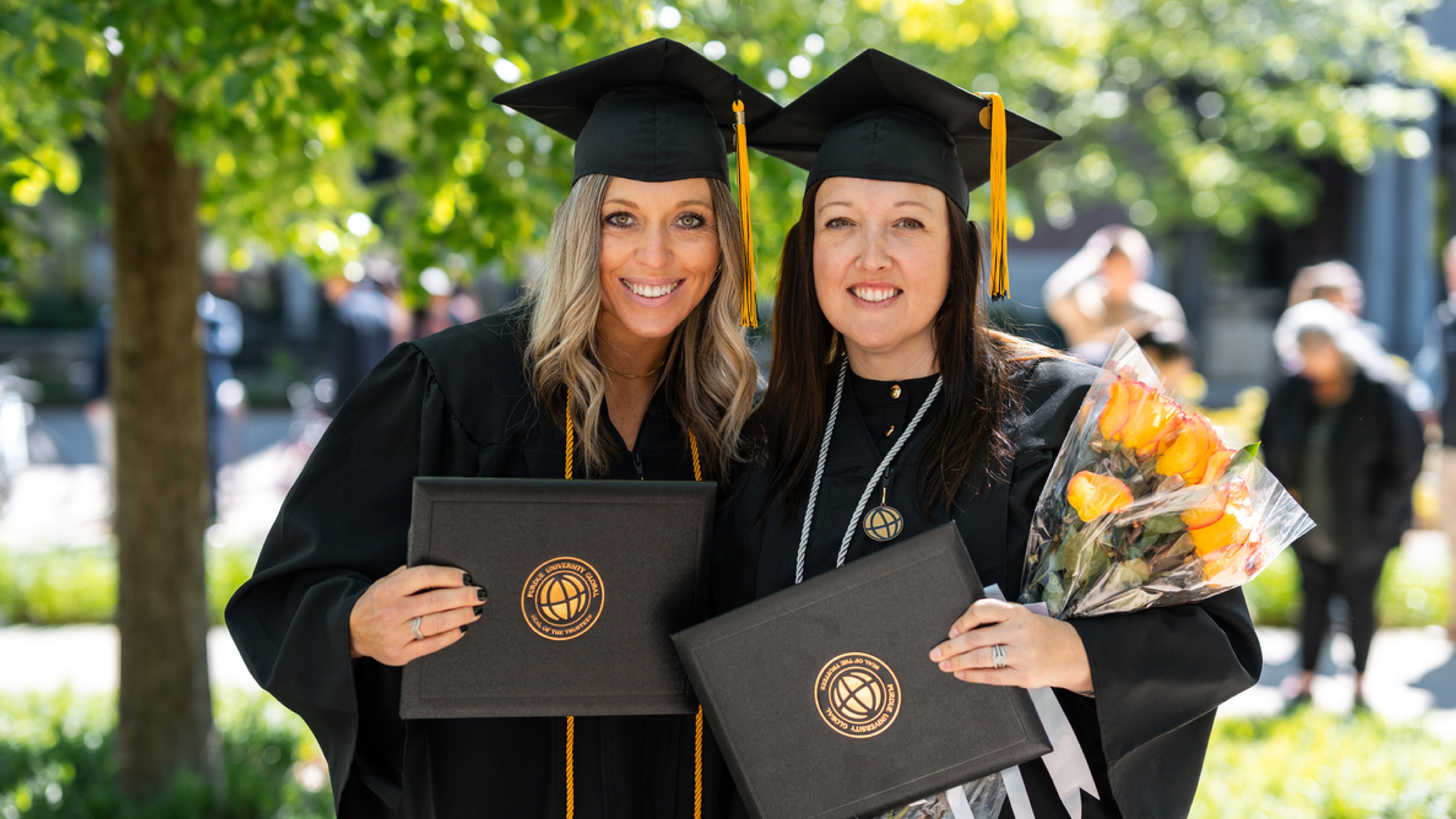cap: Stefanie Robertson (left) and Christina Walters (right) celebrate their bachelor’s degrees in healthcare administration at their in-person Purdue Global graduation ceremony. (Purdue Global photo/Kelsey Lefever)
alt: Stefanie Robertson and Christina Walters stand next to each other holding their degrees.
