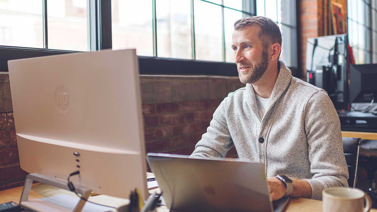 Man in beige sweater in modern office working on a laptop with a monitor