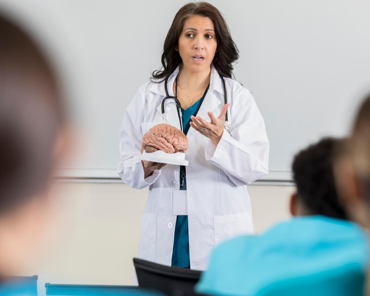 Nurse educator wearing a lab coat and stethoscope stands in front of a whiteboard while speaking to nurses in scrubs
