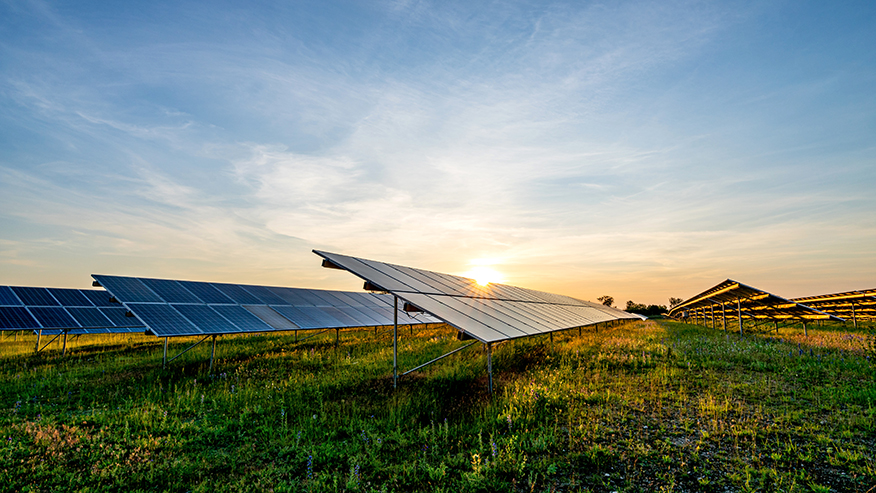 Solar panels in a grassy field at sunset, with sun rays peeking through the panels against a blue and orange sky