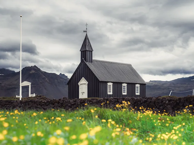 Budir, Snæfellsnes, Island Schwarze Kirche umgeben von Wiesen mit gelben Blumen und Bergen unter wolkigem Himmel, Budir, Island