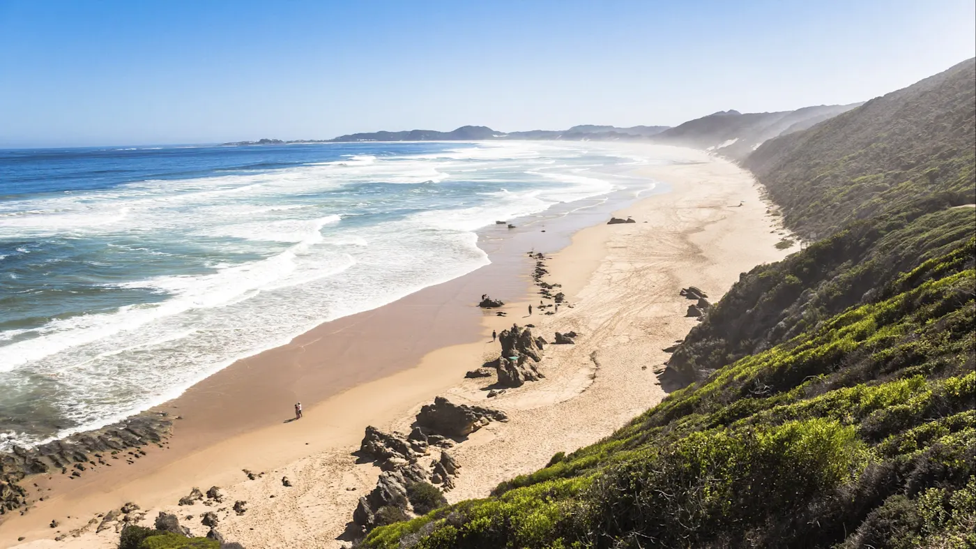 Mossel Bay, Westkap, Südafrika Langer Sandstrand mit felsiger Küste und grünen Hügeln am Ozean. Mossel Bay, Westkap, Südafrika.