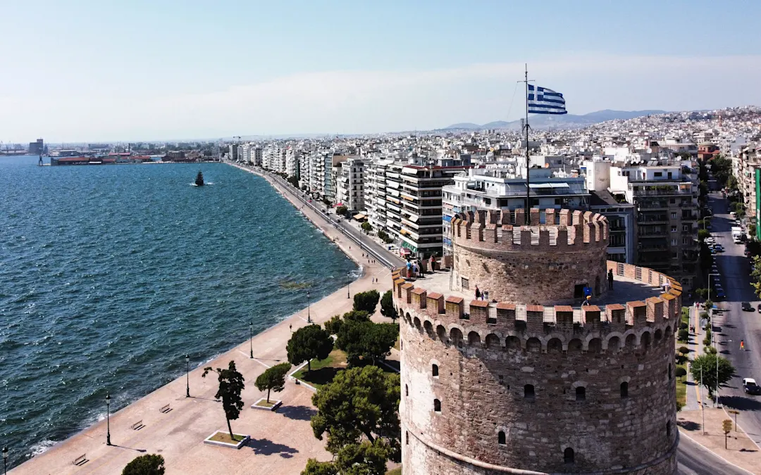 Luftaufnahme von Thessaloniki mit dem Weißen Turm, griechischer Flagge und Küstenpromenade am blauen Meer.