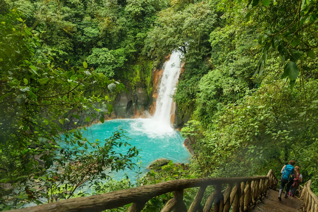 Costa Rica, Wasserfall Wasserfall fließt in ein natürliches Becken mitten im Regenwald, rechts davon Wanderer auf einer Holztreppe
