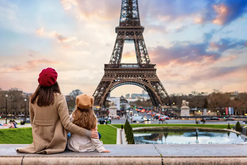 Person in red beret and child in tan hat sitting together, admiring Eiffel Tower at sunset with colorful sky.