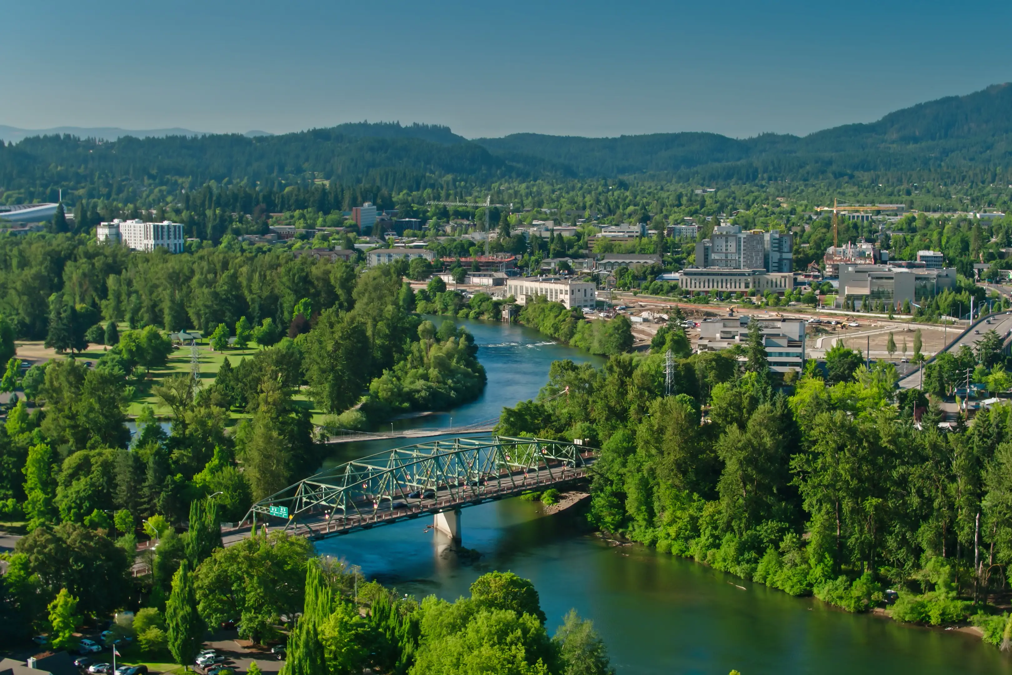 Grüne Stadtlandschaft mit Fluss, Brücke und bewaldeten Hügeln im Hintergrund, Eugene, Oregon, USA. 