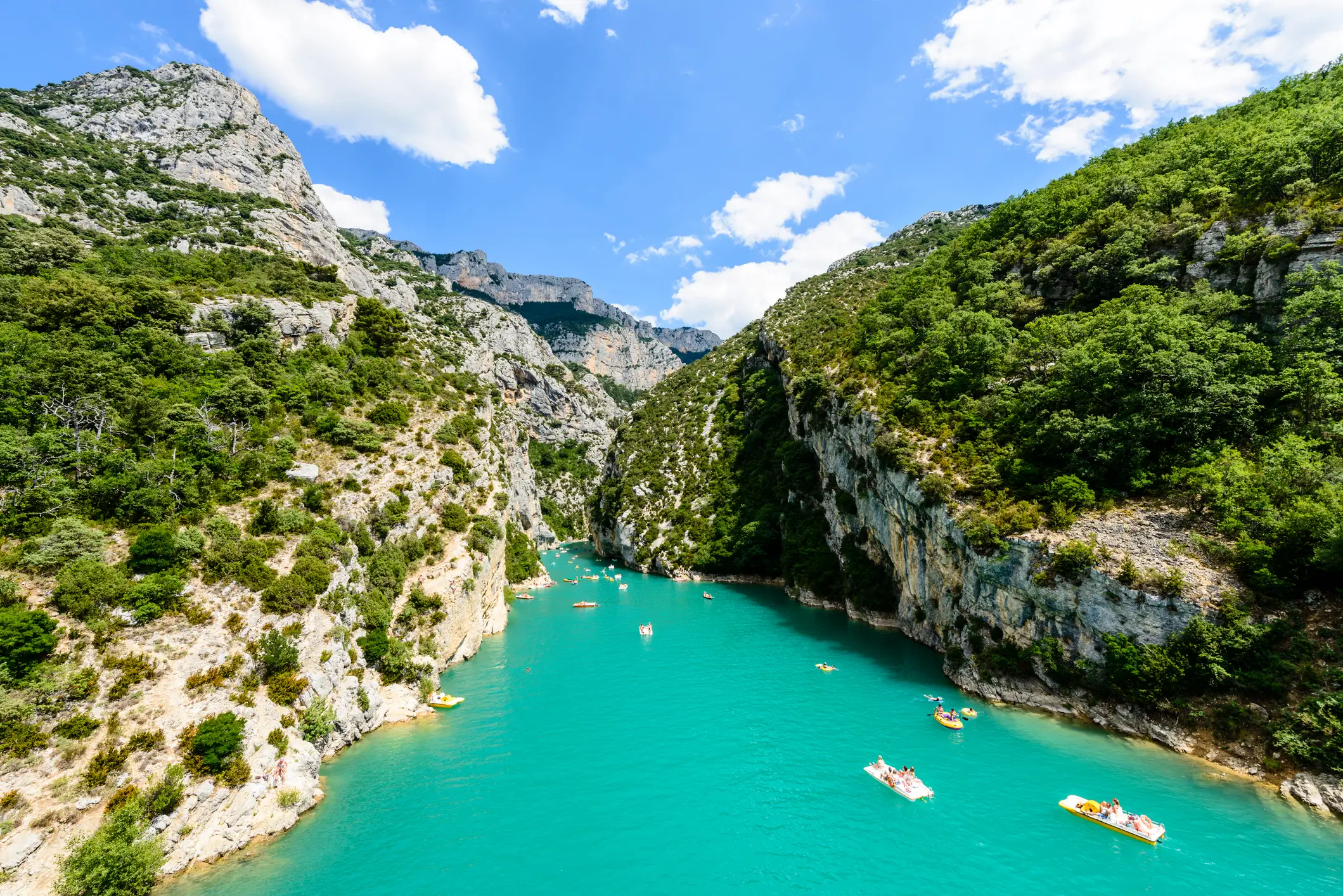 Ein türkisfarbener Fluss fließt zwischen steilen, felsigen Klippen, die mit grünen Bäumen bewachsen sind. Mehrere kleine Boote und Kajaks treiben auf dem Wasser unter einem strahlend blauen Himmel mit vereinzelten Wolken.