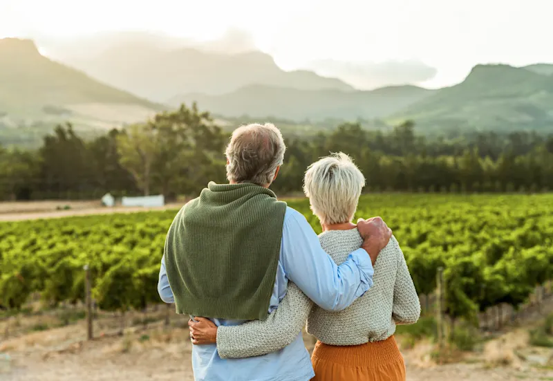 Elderly couple embracing while gazing at mountain landscape with vineyard in foreground at sunset.