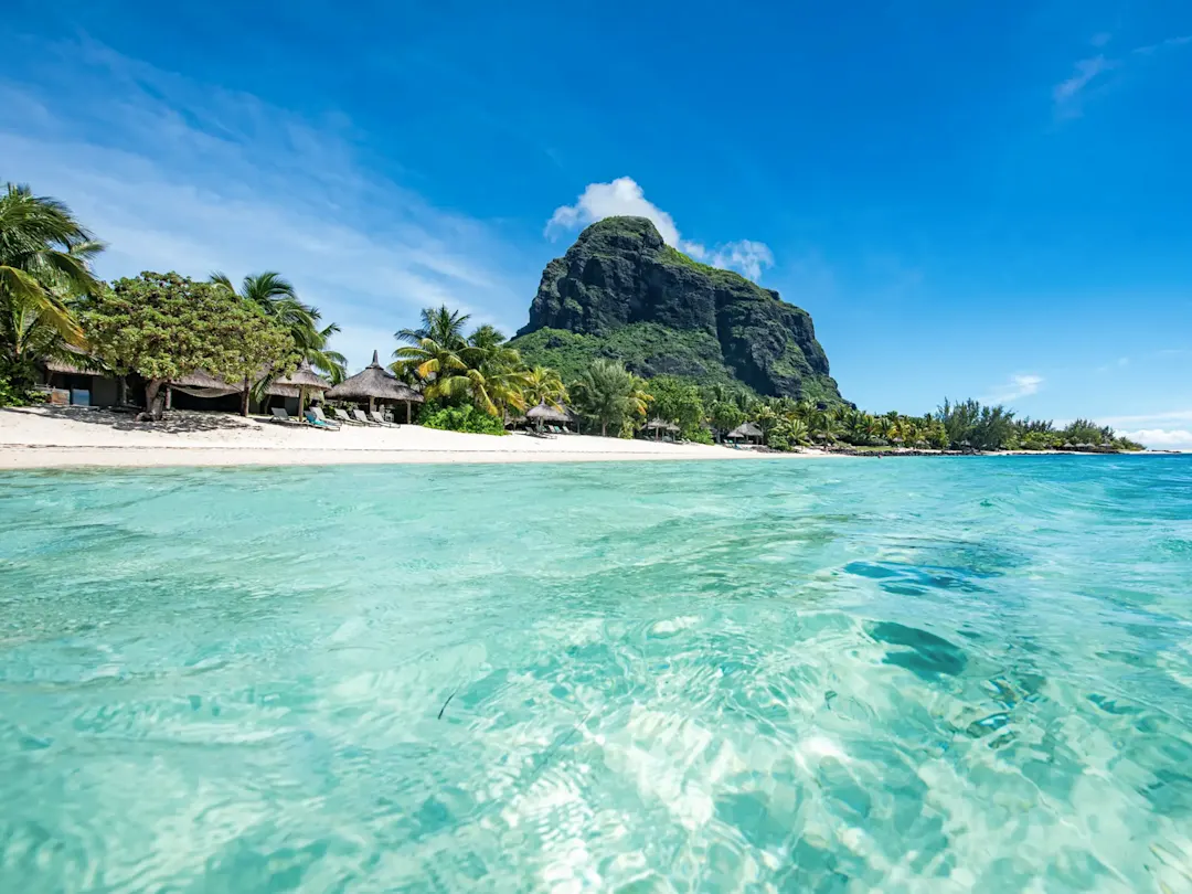 Tropischer Strand mit türkisfarbenem Wasser, Strohhütten, Palmen und Berg. Mauritius.