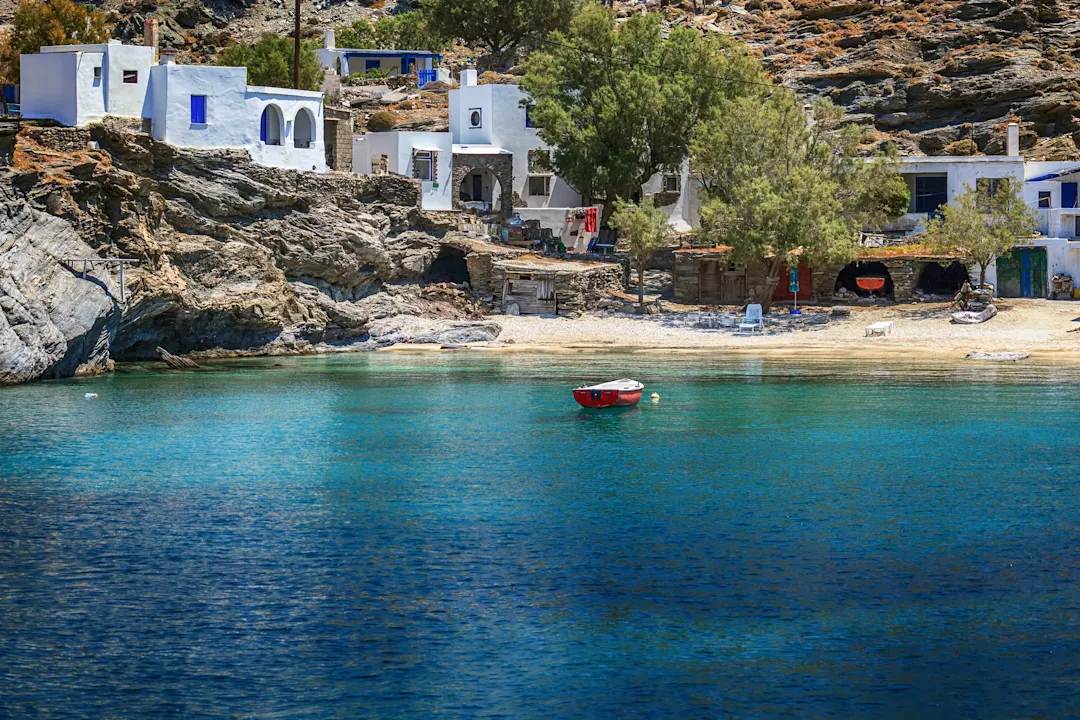 Typisch griechische Küstenlandschaft mit weißen Häusern an felsiger Bucht, türkisblauem Wasser und einem kleinen roten Boot.