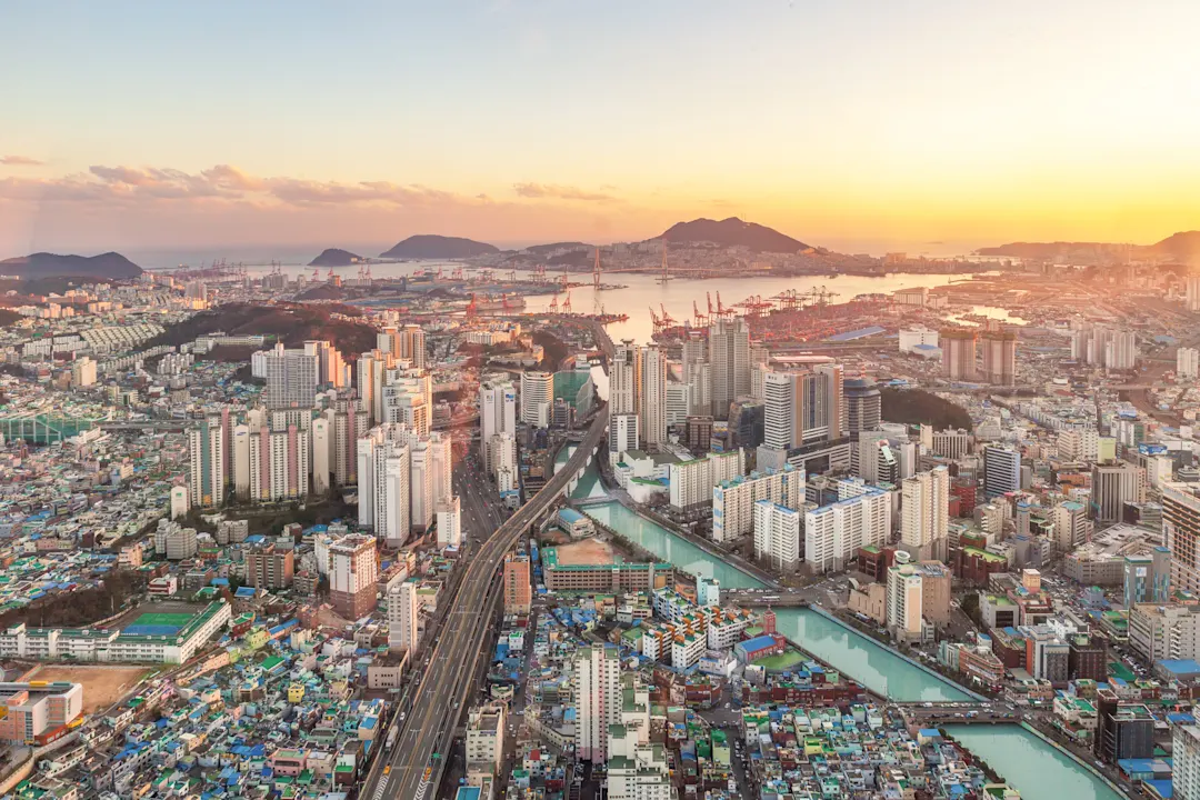 Luftaufnahme der Stadt Busan bei Sonnenuntergang. Die Sonne taucht die Stadt in ein sanftes Licht, man sieht die Küste im Hintergrund und den Kanal sowie Hochhäuser und Straßen.