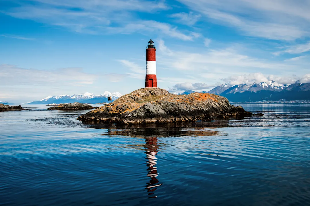 Roter und weißer Leuchtturm auf einer kleinen Insel, umgeben von Wasser und Bergen. Ushuaia, Tierra del Fuego, Argentinien.