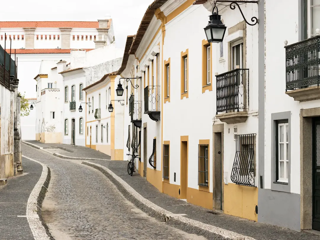 Kopfsteinpflasterstraße mit weißen Gebäuden, Balkonen und traditioneller Straßenbeleuchtung. Evora, Portugal.