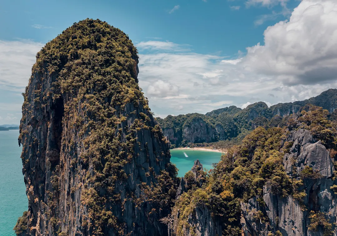 Dramatische Karstfelsen mit üppiger Vegetation umgeben eine türkisfarbene Bucht mit kleinem Strand unter blauem Himmel.
