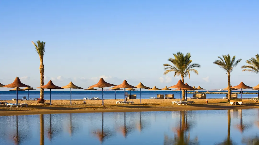 Ein ruhiger Strand mit Sonnenschirmen und Palmen, die sich im blauen Wasser spiegeln, unter wolkenlosem Himmel, in Hurghada, Ägypten.