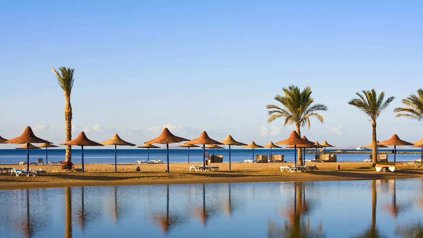 Strand, Hurghada, Ägypten Ein ruhiger Strand mit Sonnenschirmen und Palmen, die sich im blauen Wasser spiegeln, unter wolkenlosem Himmel, in Hurghada, Ägypten.