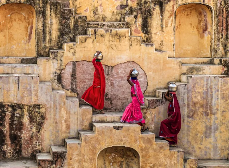 Jaipur, Rajasthan, Indien Drei indische Frauen in roten Saris tragen Wasserkrüge in einem historischen Stufenbrunnen. Jaipur, Rajasthan, Indien.