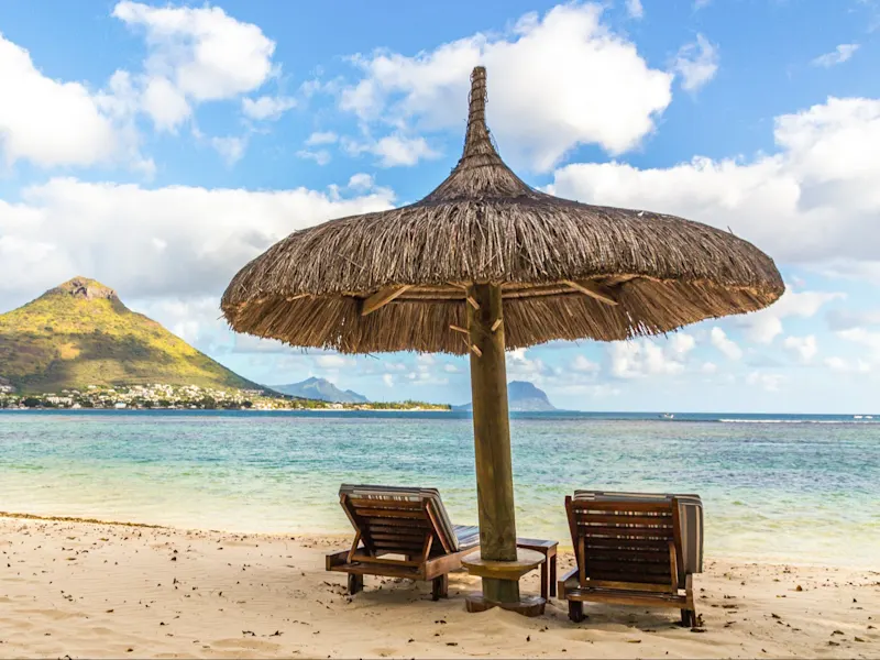 Flic en Flac, Mauritius Strand mit Strohdach-Sonnenschirm und Liegen vor Bergkulisse. Flic en Flac, Mauritius.