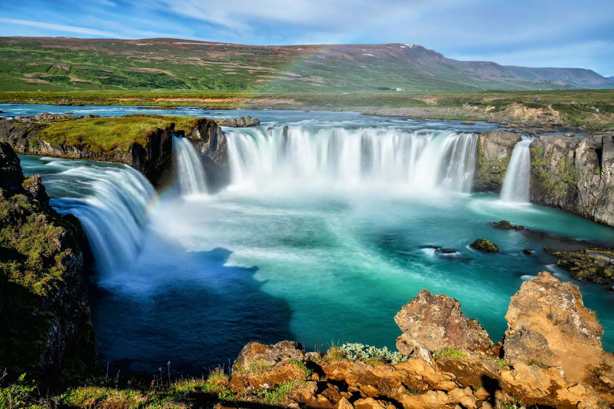 Ein breiter, mächtiger Wasserfall stürzt in einen türkisfarbenen Fluss, der von felsigen Klippen und grünen Hügeln umgeben ist. Ein schwacher Regenbogen erscheint im Nebel über dem Wasser unter einem teilweise bewölkten Himmel.