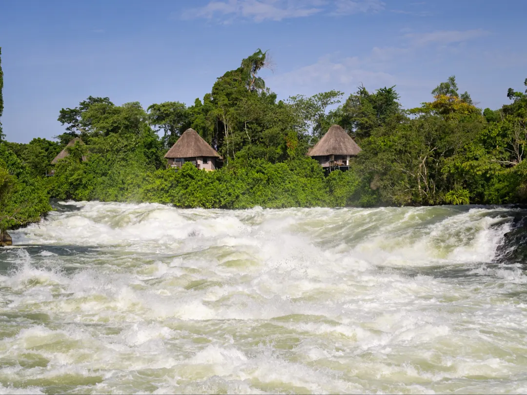 Itanda-Wasserfälle, Osten, Uganda Rauschende Stromschnellen mit grüner Vegetation und strohgedeckten Hütten. Itanda-Wasserfälle, Osten, Uganda.