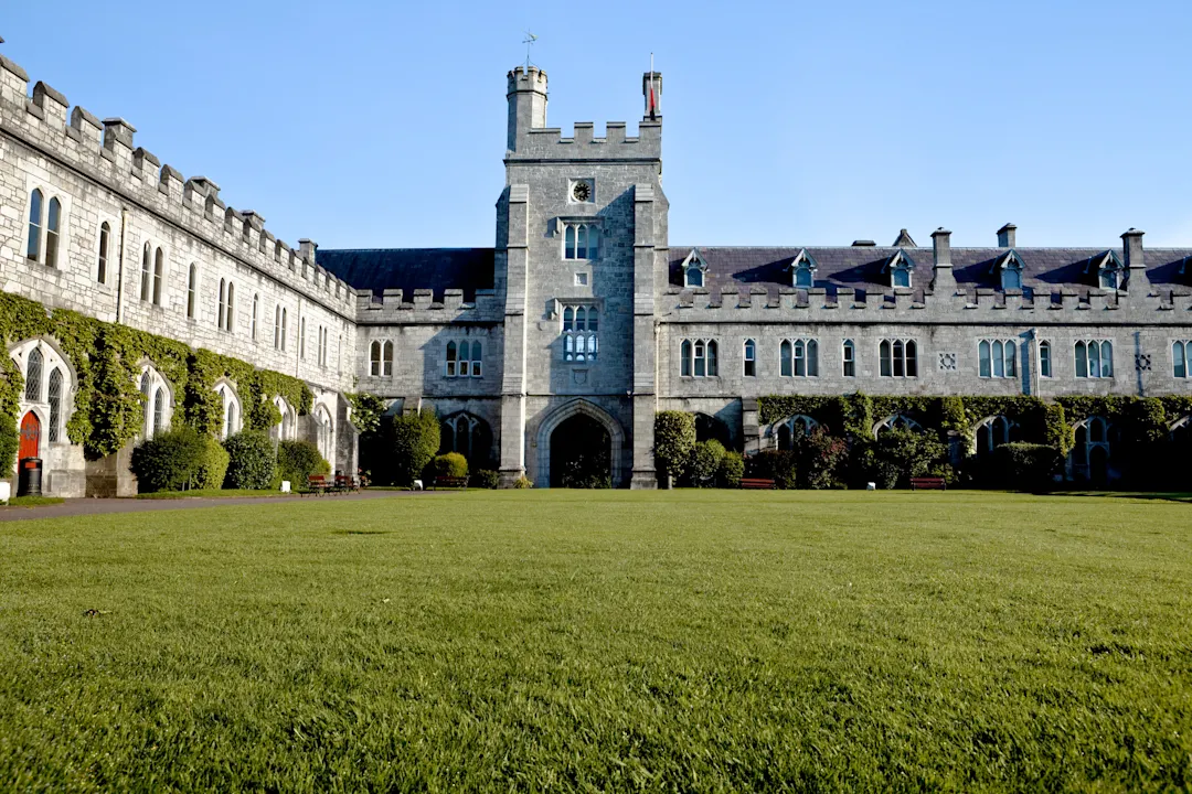 Ein großes, historisches Steingebäude mit Rundbogenfenstern und einem zentralen Turm, umgeben von grünen Rasenflächen und Sträuchern, unter einem klaren blauen Himmel.