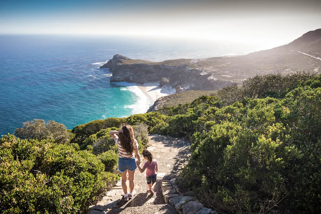 Zwei Personen wandern auf einem Küstenpfad mit Blick auf türkisblaues Meer, felsige Klippen und einen weißen Sandstrand.