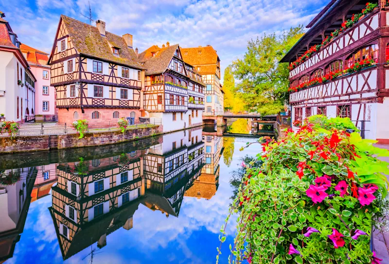Fachwerkhäuser entlang eines Kanals in Straßburg mit bunten Blumen und perfekter Spiegelung im ruhigen Wasser.