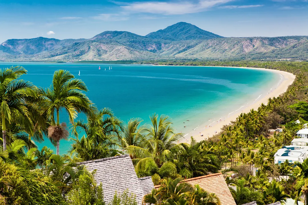 Landschaftliche Ansicht eines tropischen Strandes mit türkisfarbenem Wasser, weißem Sand, Palmen und üppig grünen Bergen im Hintergrund unter blauem Himmel. In der Ferne sind Segelboote zu sehen.