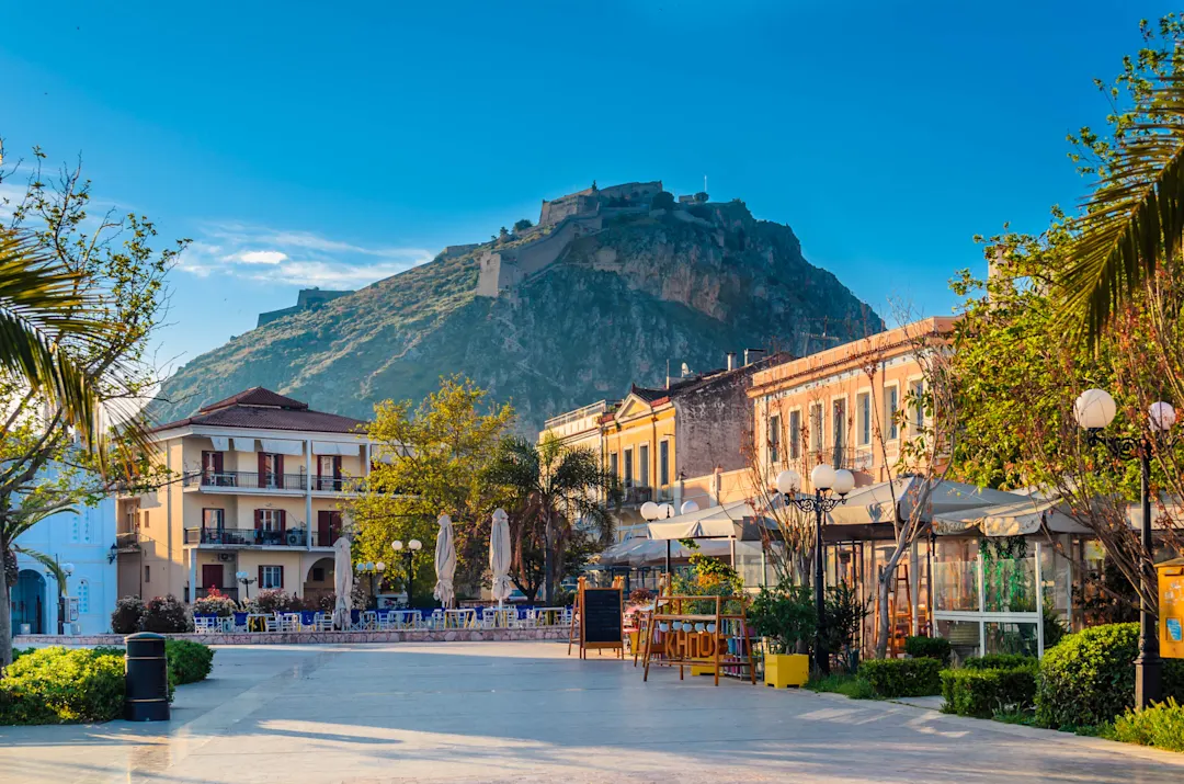 Malerische griechische Stadt mit bunten Gebäuden und Cafés unter einer imposanten Festung auf einem Berg bei blauem Himmel.