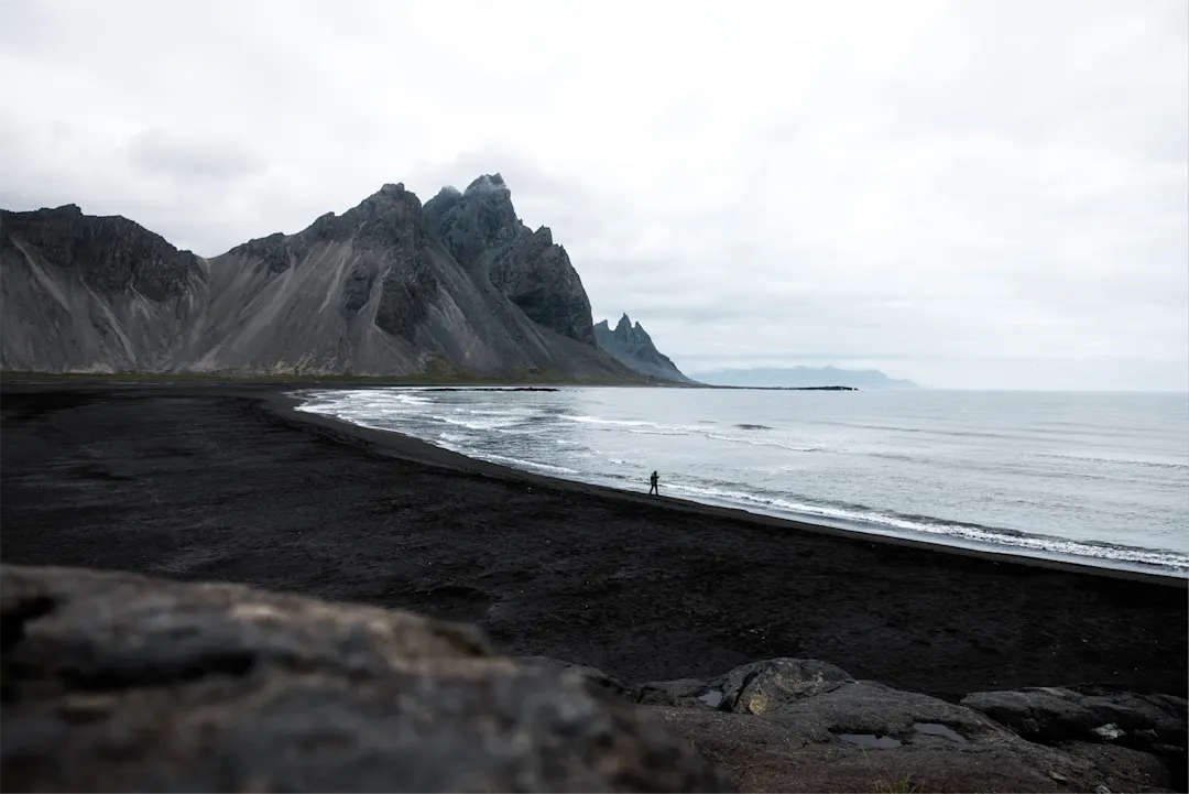 Dramatische Küstenlandschaft mit schwarzem Strand und schroffen Bergen in Island, eine einsame Person am Ufer.