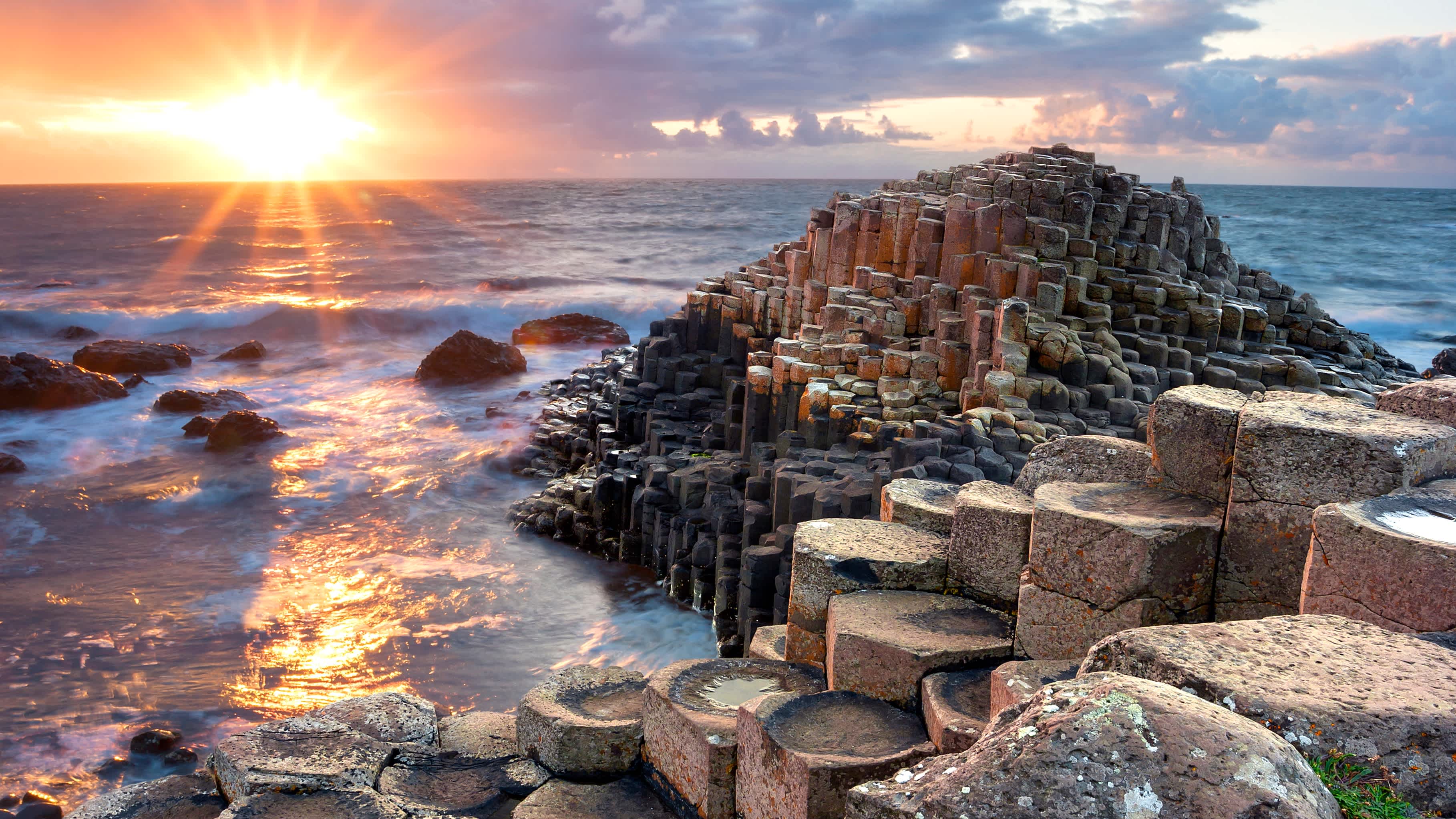 Discover the Giant's Causeway on a Game of Thrones Ireland tour. The sky is purple and the water breaks on the basalt columns. 