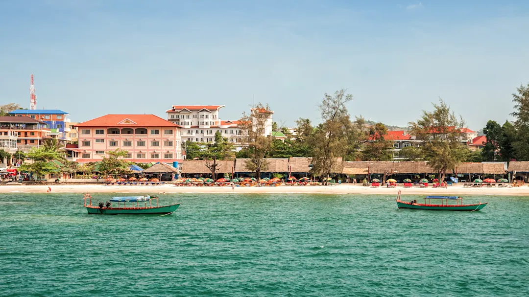 Boote auf türkisfarbenem Wasser vor einem Sandstrand mit Hotels. Sihanoukville, Kambodscha.
