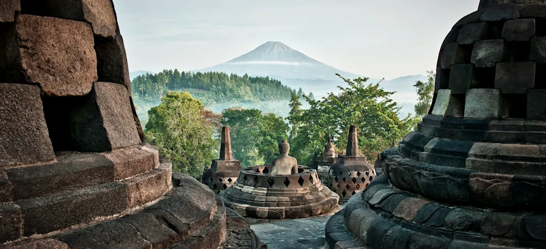 Blick vom Borobudur-Tempel auf Buddha-Statuen in Stupas mit dem majestätischen Vulkan Merapi im Hintergrund.