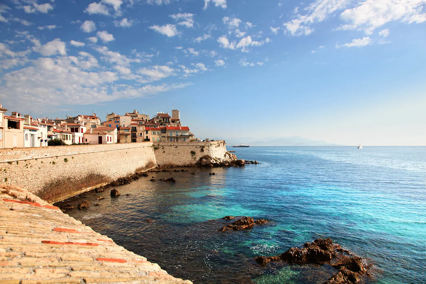 Coastal Mediterranean town with white and terracotta buildings perched on stone walls above turquoise waters under blue skies.
