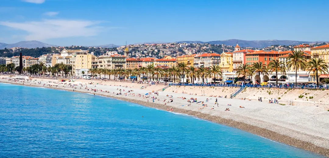 Panoramablick auf den Strand von Nizza mit türkisblauem Meer, Kieselstrand, Palmenpromenade und bunten Gebäuden.