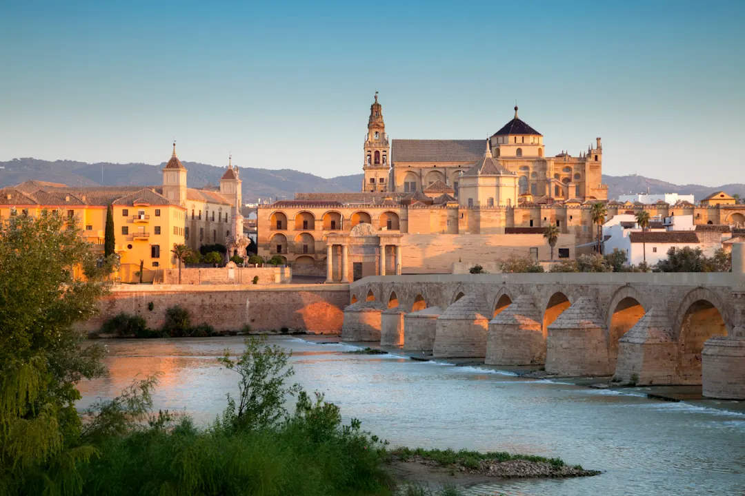 Blick auf die Römische Brücke und die Moschee-Kathedrale von Córdoba bei Sonnenuntergang, mit Steinbögen, die den Fluss überqueren, und historischen Gebäuden unter einem klaren blauen Himmel.