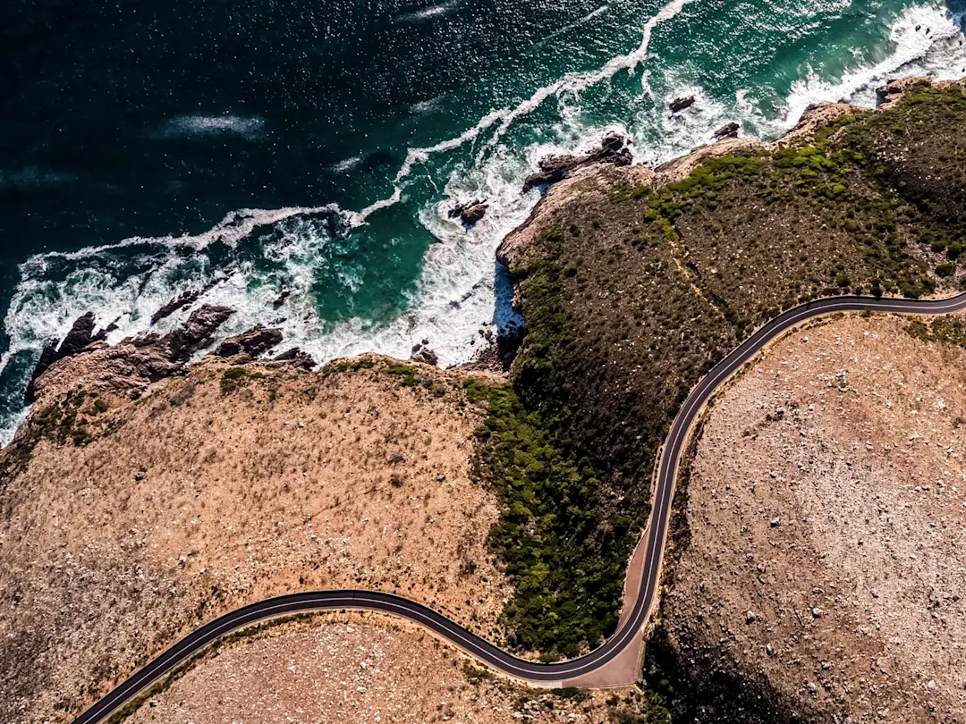 Luftaufnahme einer kurvigen Küstenstraße entlang rauer Klippen und türkisfarbenem Wasser. Südafrika.