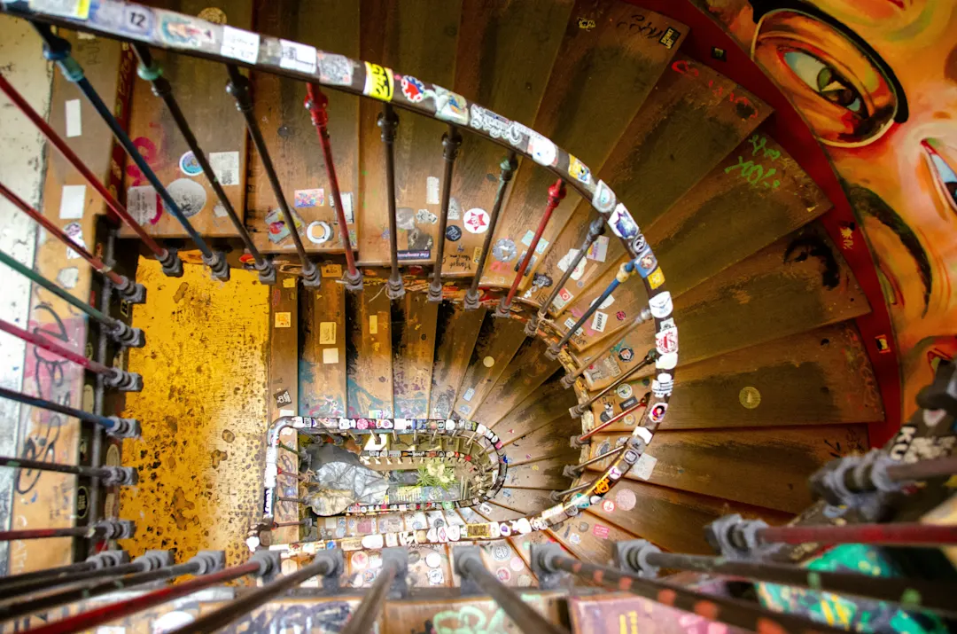 Spiralförmige Holztreppe von oben fotografiert, mit Aufklebern verziert und Graffiti an den Wänden, gelber Boden unten sichtbar.