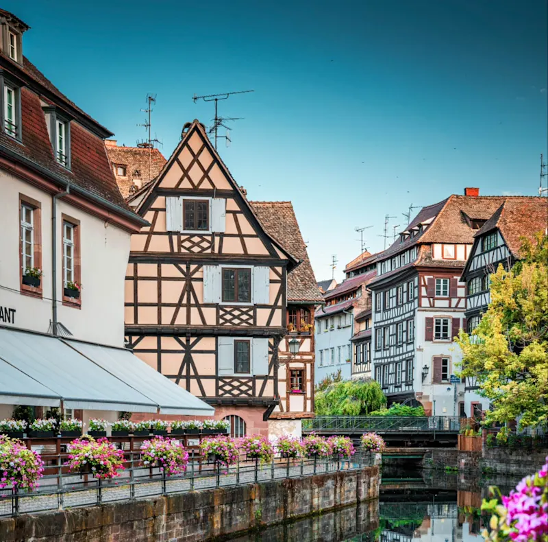 Colorful half-timbered buildings along a canal in Strasbourg with flower boxes and clear blue sky.