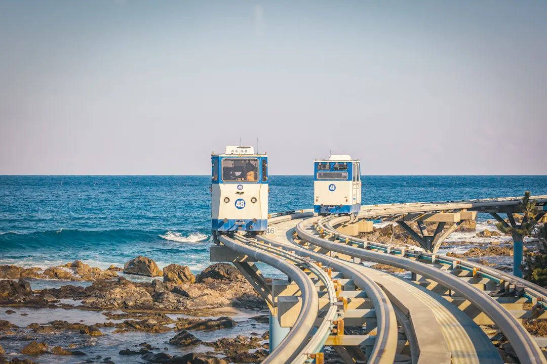 Zwei blau-weiße Küstenbahnen fahren auf geschwungenen Schienen über felsiger Meeresküste bei klarem Himmel.