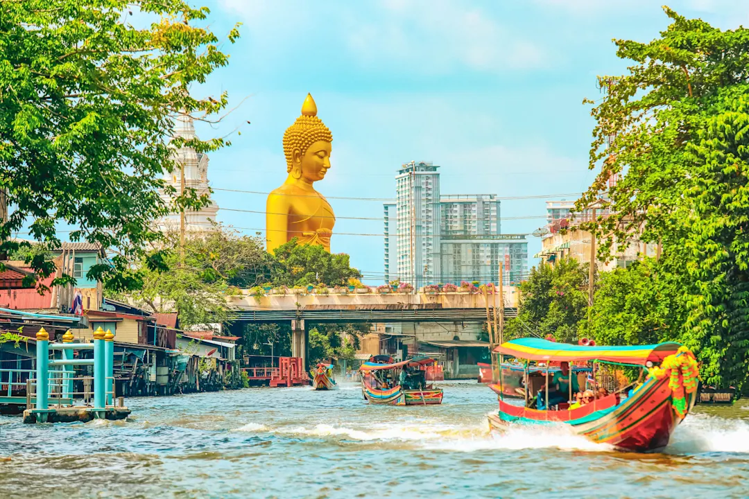 Goldene Buddha-Statue überragt einen Kanal in Bangkok mit bunten Booten und modernen Gebäuden im Hintergrund.