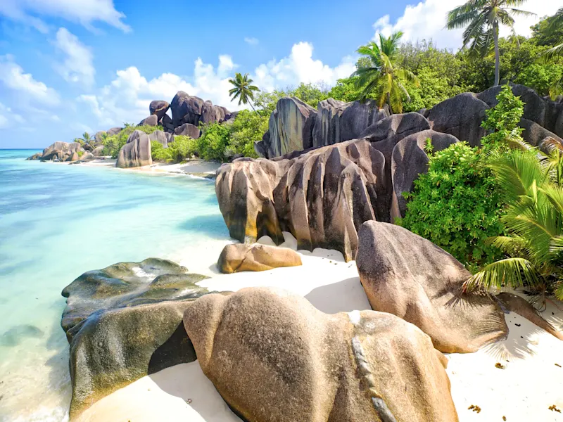 Plage avec rochers granitiques, eau turquoise, ciel bleu, palmiers, La Digue, Seychelles.