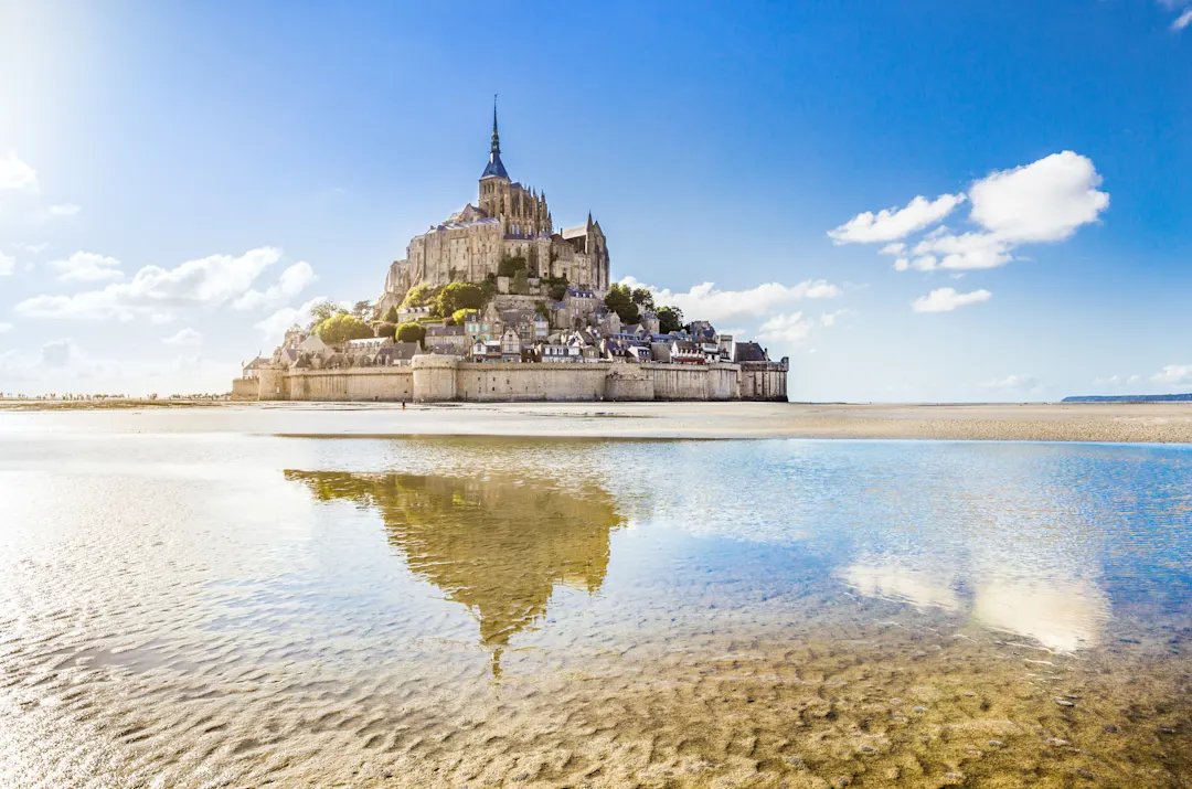 Der Mont Saint-Michel, eine historische Inselgemeinde in Frankreich, erhebt sich mit seiner ikonischen Abtei und seinen mittelalterlichen Gebäuden über dem sandigen Wattenmeer und spiegelt sich im seichten Wasser unter einem strahlend blauen Himmel mit vereinzelten Wolken.