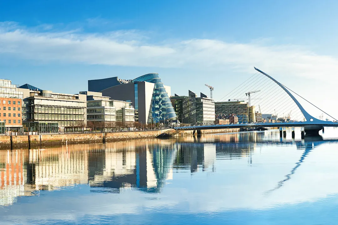 Moderne Gebäude und Büros am Liffey River in Dublin an einem hellen, sonnigen Tag, Brücke auf der rechten Seite ist eine berühmte Harfenbrücke.