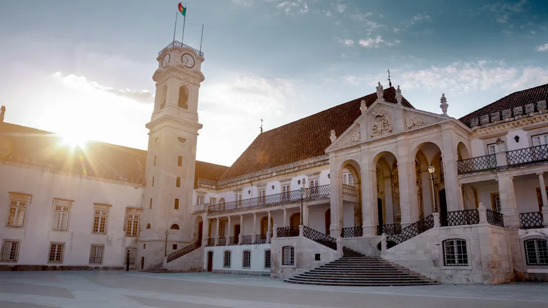 Das Bild zeigt das historische Gebäude der Universität von Coimbra in Portugal mit einem Uhrenturm, einem gewölbten Eingang, einer verzierten Fassade und der strahlenden Sonne im Hintergrund.