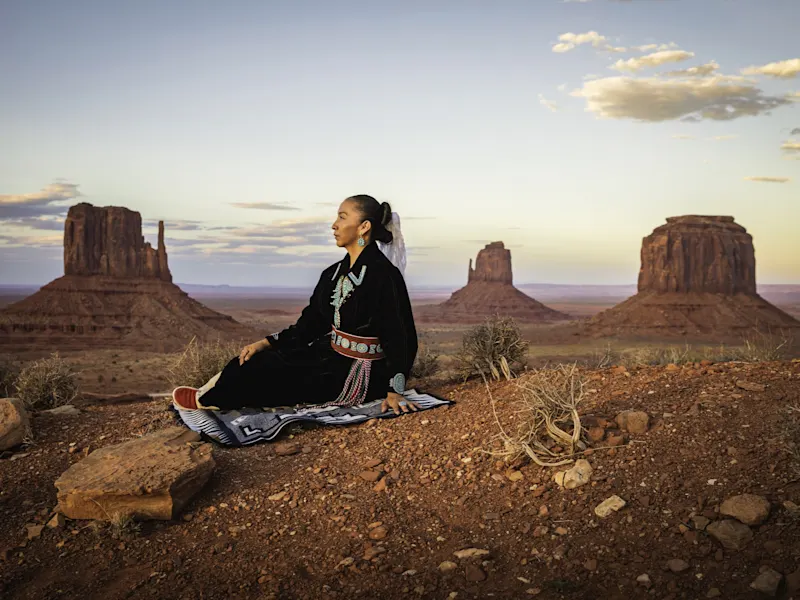 Frau in traditioneller Kleidung sitzt vor den Felsformationen im Monument Valley. Monument Valley, Arizona, USA.