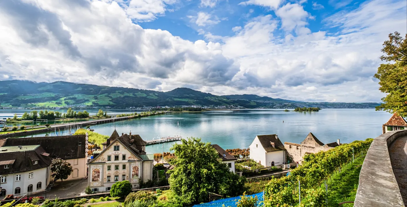 Scenic lakeside village with historic buildings nestled beneath dramatic blue sky and mountains, overlooking calm waters.
