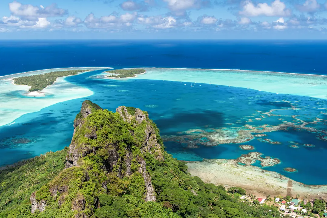 Luftaufnahme einer tropischen Insel mit grünem Felsen im Vordergrund, türkisfarbener Lagune und Korallenriff im blauen Ozean.