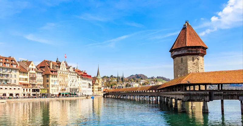 Historic Chapel Bridge and Water Tower in Lucerne, Switzerland, with colorful buildings along the waterfront under blue sky.
