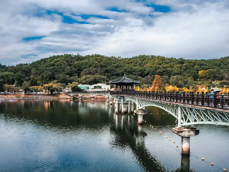 Südkorea, Andong, Roadtrip Brücke mit traditionellem Pavillon, reflektierendes Wasser, grüne Hügel, teilweise bewölkter Himmel. Andong, Korea.